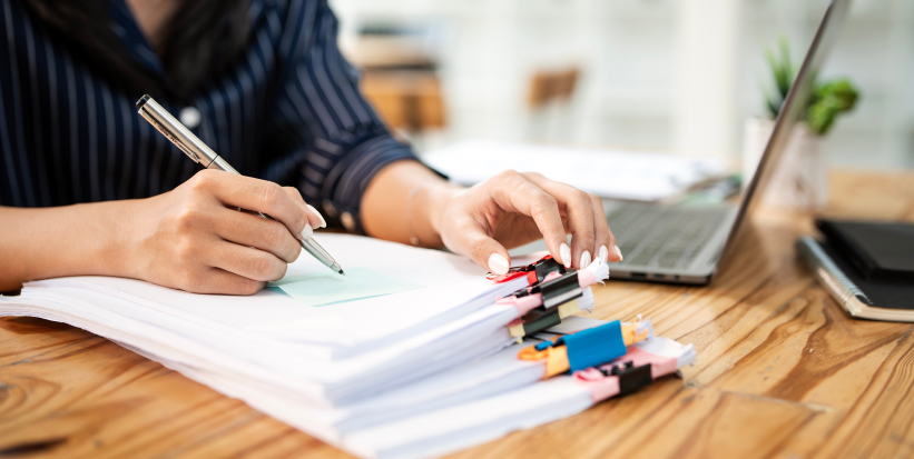 A stack of paper files sits on a desk in an office setting. An office worker writes a post-it note on the top file.